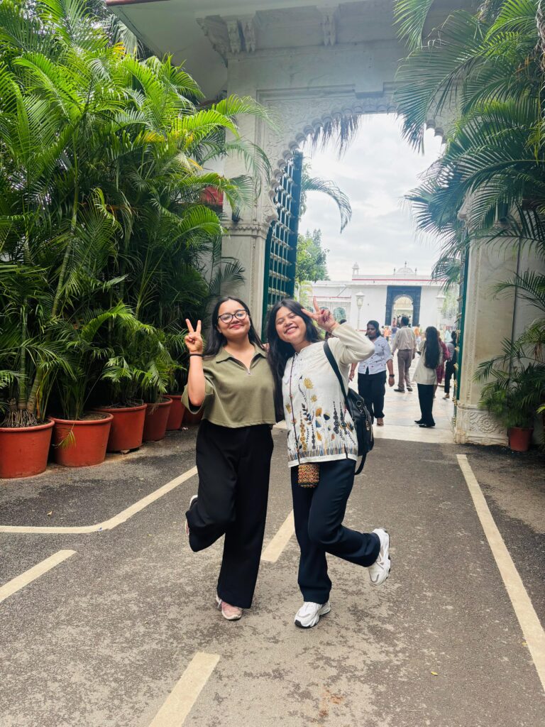 Two friends striking a playful pose on a palm-lined path leading to a grand palace gateway